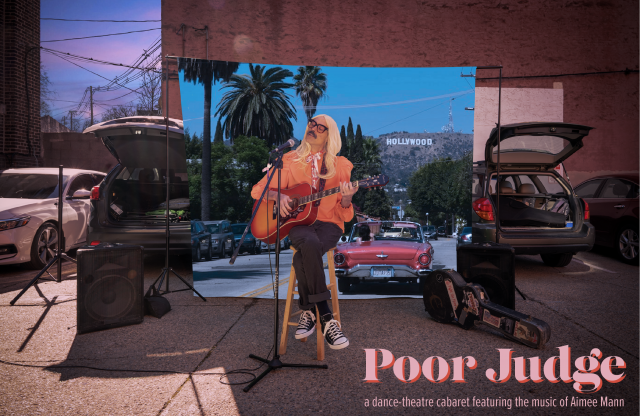Dito dressed as Aimee Mann sits in a parking lot with an image of LA behind him